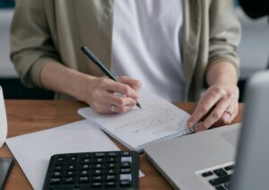 A woman writes financial calculations in a notebook, using a calculator and laptop at a wooden desk.